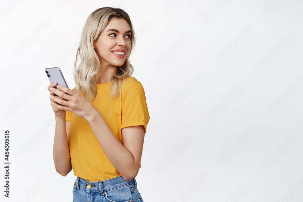 Celluar technology. Young blond woman holds mobile phone, turn head behind and smile at copy space, stands in yellow tshirt over white background Celluar technology. Young blond woman holds mobile phone, turn head behind and smile at copy space, stands in yellow tshirt over white background