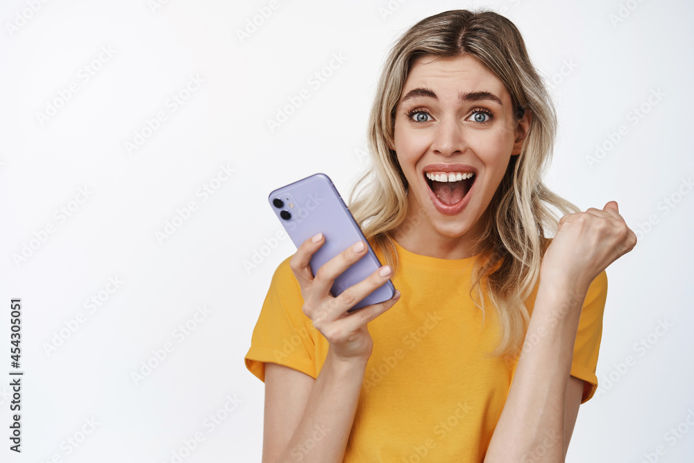 Close up portrait of excited woman winning on mobile phone, holding smartphone and cheering, triumphing achieve goal, standing over white background Close up portrait of excited woman winning on mobile phone, holding smartphone and cheering, triumphing achieve goal, standing over white background