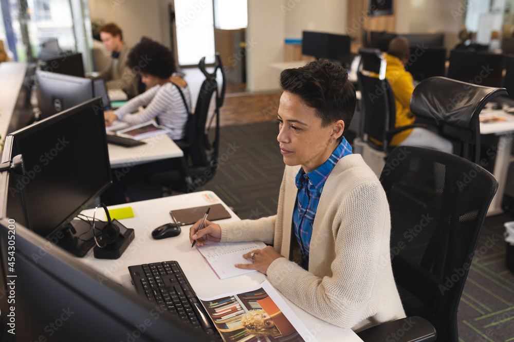 Mixed race female creative at work, sitting at desk, using computer Mixed race female creative at work, sitting at desk, using computer
