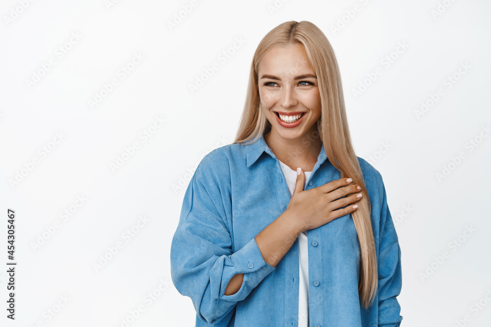 Beautiful female model holding hand on heart and smiling, looking left at copy space, thank you gesture, standing over white background Beautiful female model holding hand on heart and smiling, looking left at copy space, thank you gesture, standing over white background