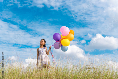 Cheerful cute girl holding balloons running on green meadow white cloud and blue sky with happiness. Hands holding vibrant air balloons play on birthday party happy times summer on sunlight outdoor