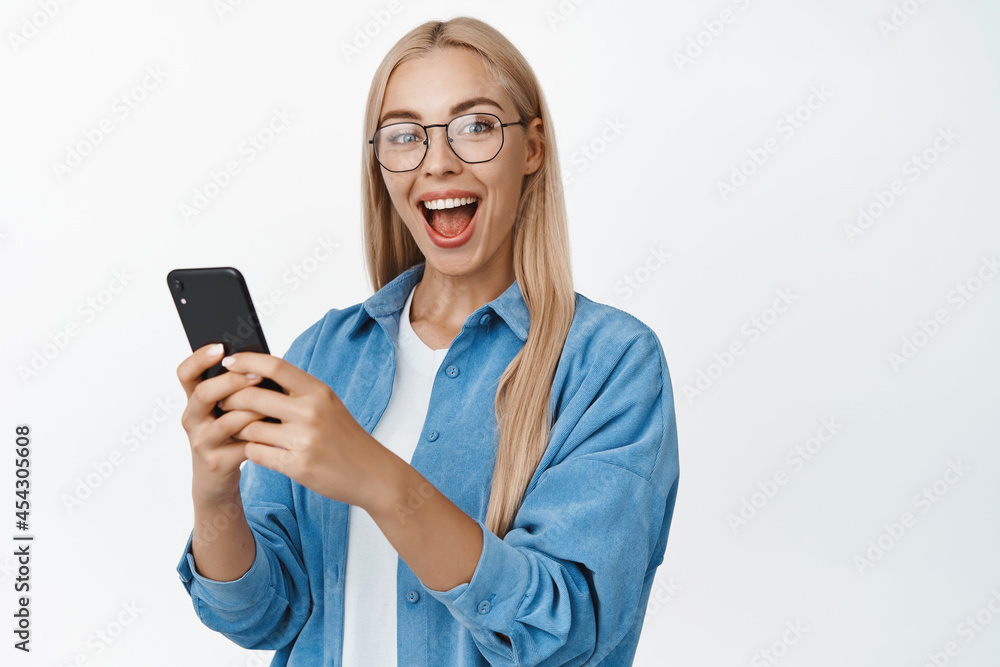 Portrait of excited smiling girl holding mobile phone and reacting amazed at camera, wearing glasses, standing over white background