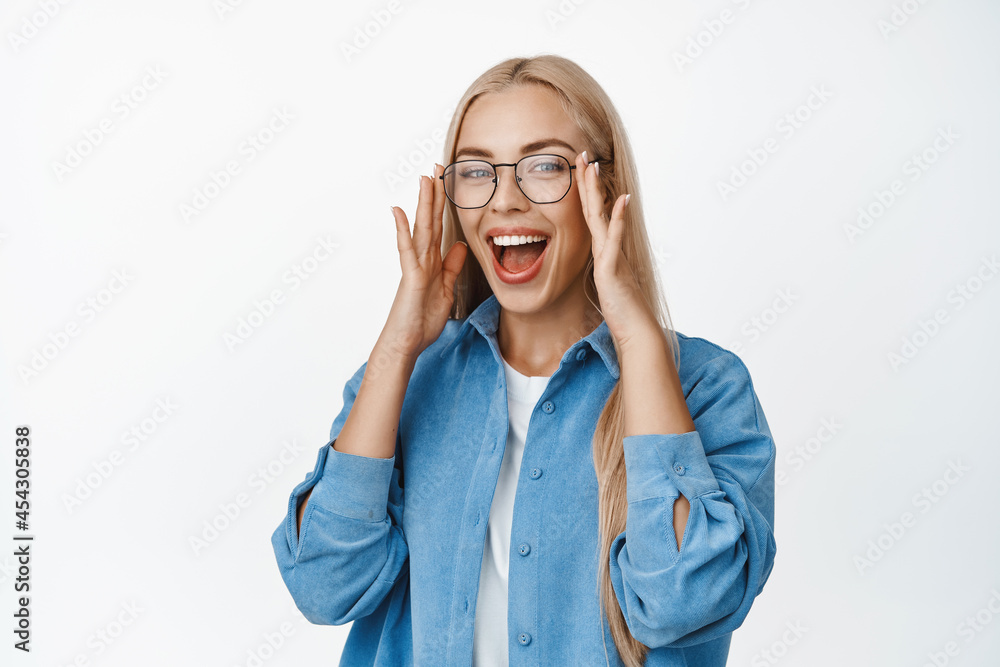 Beautiful blond woman put on glasses, laughing and smiling with happy face expression, standing in blue shirt over white background Beautiful blond woman put on glasses, laughing and smiling with happy face expression, standing in blue shirt over white background