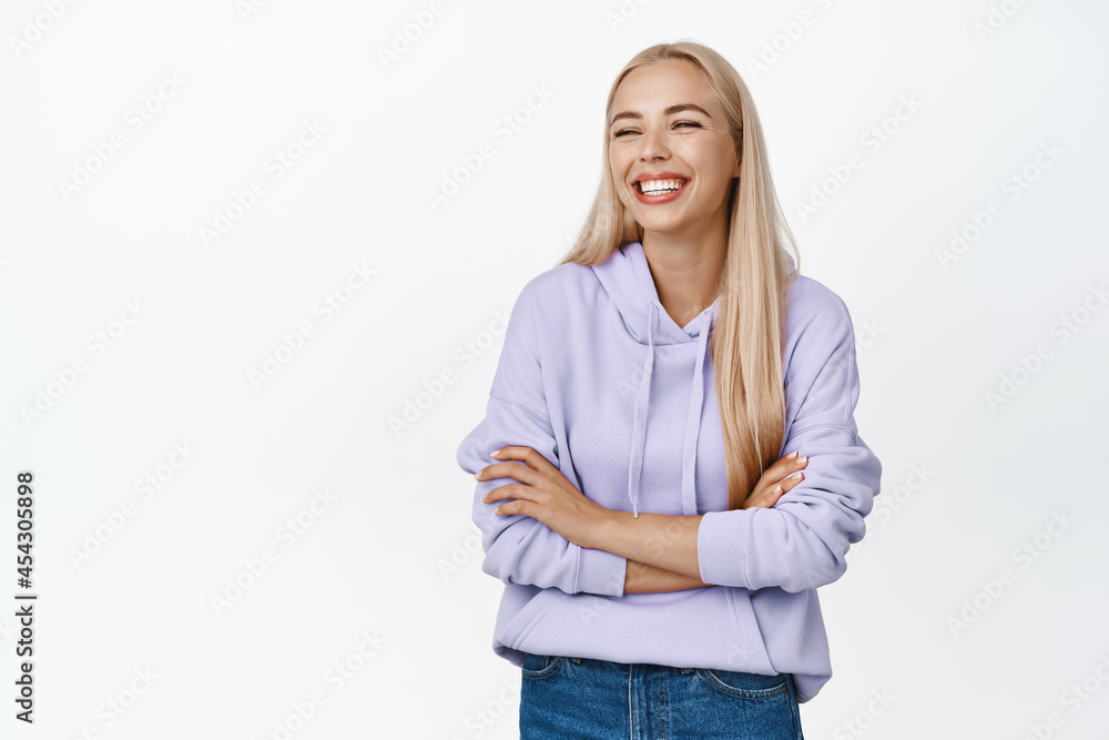Happy young woman looking aside, smiling and laughing, standing in casual relaxed pose with arms crossed over chest, white background Happy young woman looking aside, smiling and laughing, standing in casual relaxed pose with arms crossed over chest, white background