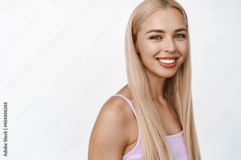 Beautiful blond girl smiling and looking happy at camera, turn her head while standing in profile in tank-top, concept of makeup and cosmetics, white background Beautiful blond girl smiling and looking happy at camera, turn her head while standing in profile in tank-top, concept of makeup and cosmetics, white background