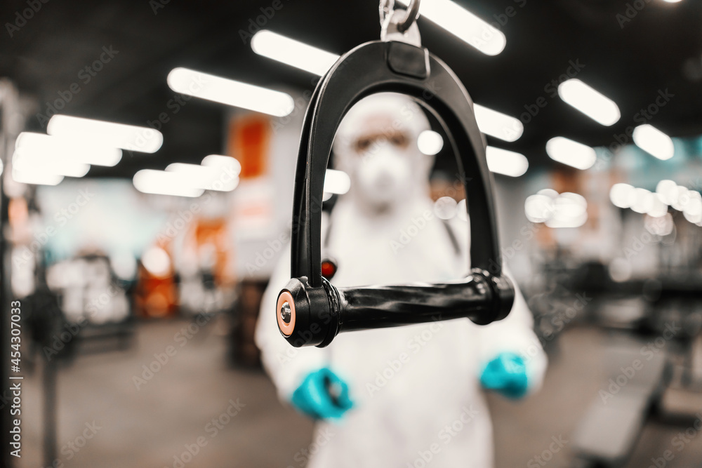 The serious situation with the corona virus. Suppression of COVID-19 virus and cleaning of exercise equipment in the gym by a male person in a protective white suit The serious situation with the corona virus. Suppression of COVID-19 virus and cleaning of exercise equipment in the gym by a male person in a protective white suit