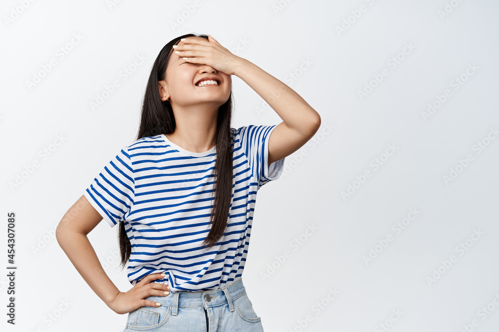Beautiful korean girl covering her eyes for surprise, smiling from anticiaption, waiting for something blindfolded, standing over white background Beautiful korean girl covering her eyes for surprise, smiling from anticiaption, waiting for something blindfolded, standing over white background