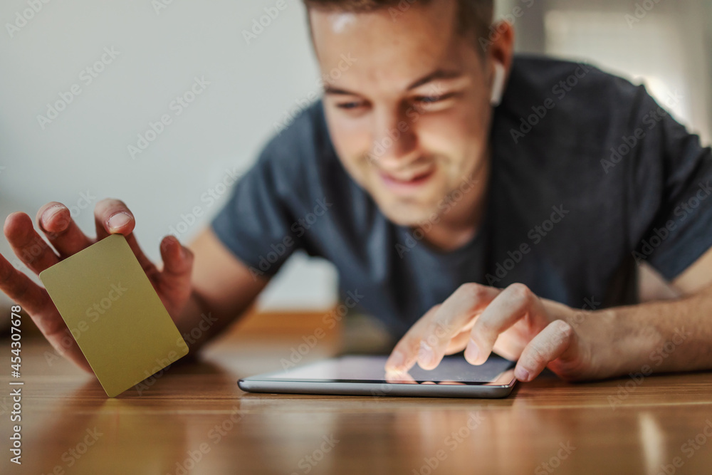Online purchase with a gold credit card on a tablet. An excited man lies on his stomach on the floor with underfloor heating and uses a tablet with a touchscreen and holds a card in the other hand Online purchase with a gold credit card on a tablet. An excited man lies on his stomach on the floor with underfloor heating and uses a tablet with a touchscreen and holds a card in the other hand