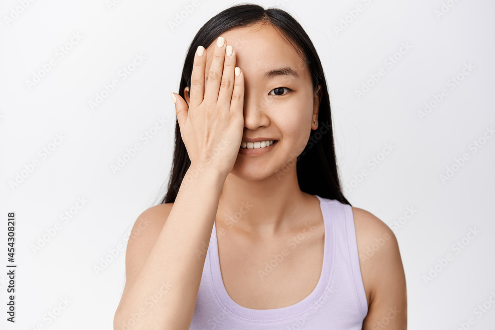 Close up portrait of cute asian girl without makeup, covers one side of face with hand, smiling and looking cheerful at camera, white background Close up portrait of cute asian girl without makeup, covers one side of face with hand, smiling and looking cheerful at camera, white background