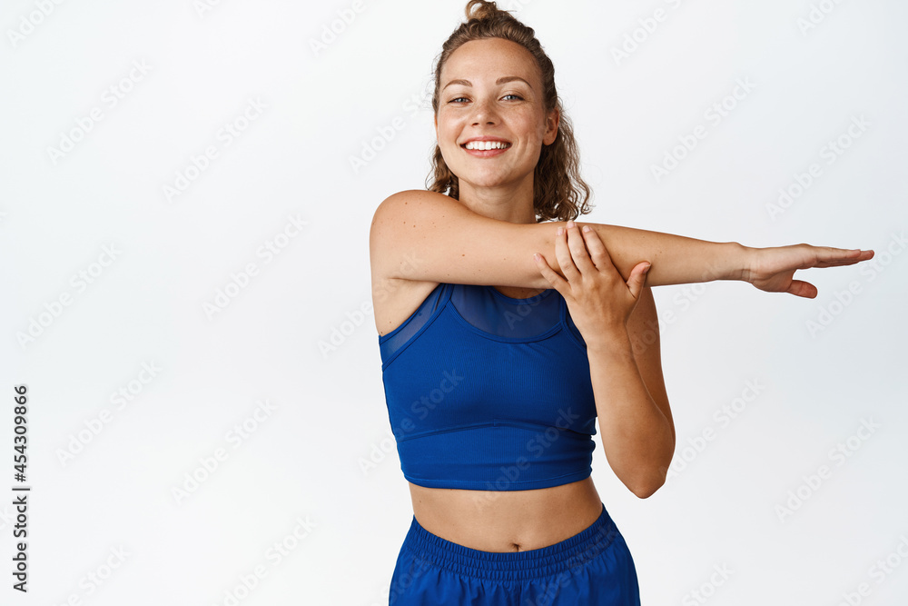 Smiling blond fitness woman stretching hands, doing exercises and workout, standing in blue sportsbra and leggings, white background Smiling blond fitness woman stretching hands, doing exercises and workout, standing in blue sportsbra and leggings, white background