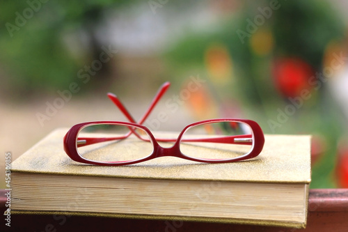 Vintage hardcover book and red reading glasses in a garden. Selective focus.