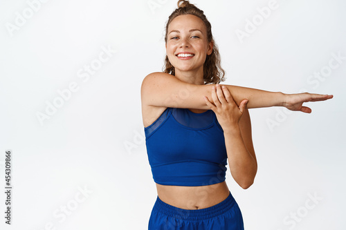 Smiling blond fitness woman stretching hands, doing exercises and workout, standing in blue sportsbra and leggings, white background