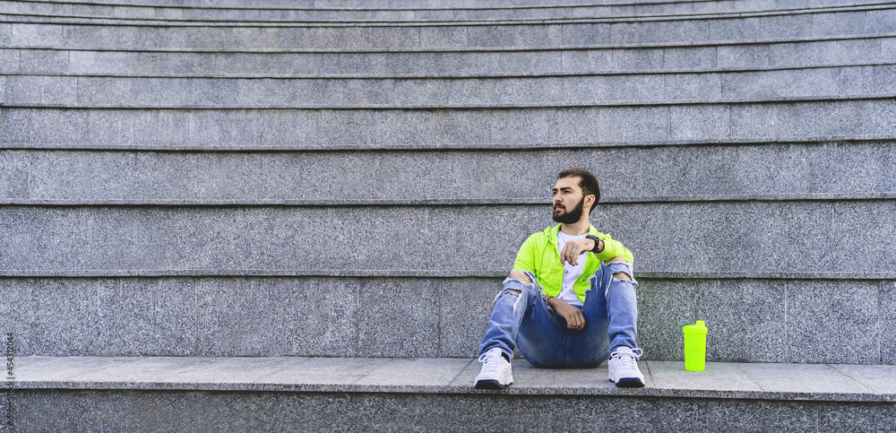Stylish hipster man with beard sits alone on the steps. Guy in trendy bright casual clothes. Banner Stylish hipster man with beard sits alone on the steps. Guy in trendy bright casual clothes. Banner