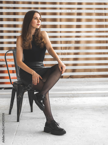 Elegant young woman in black clothes, dress, pantyhose and boots, sitting on chair in studio background. Future and urban fashion concept photo