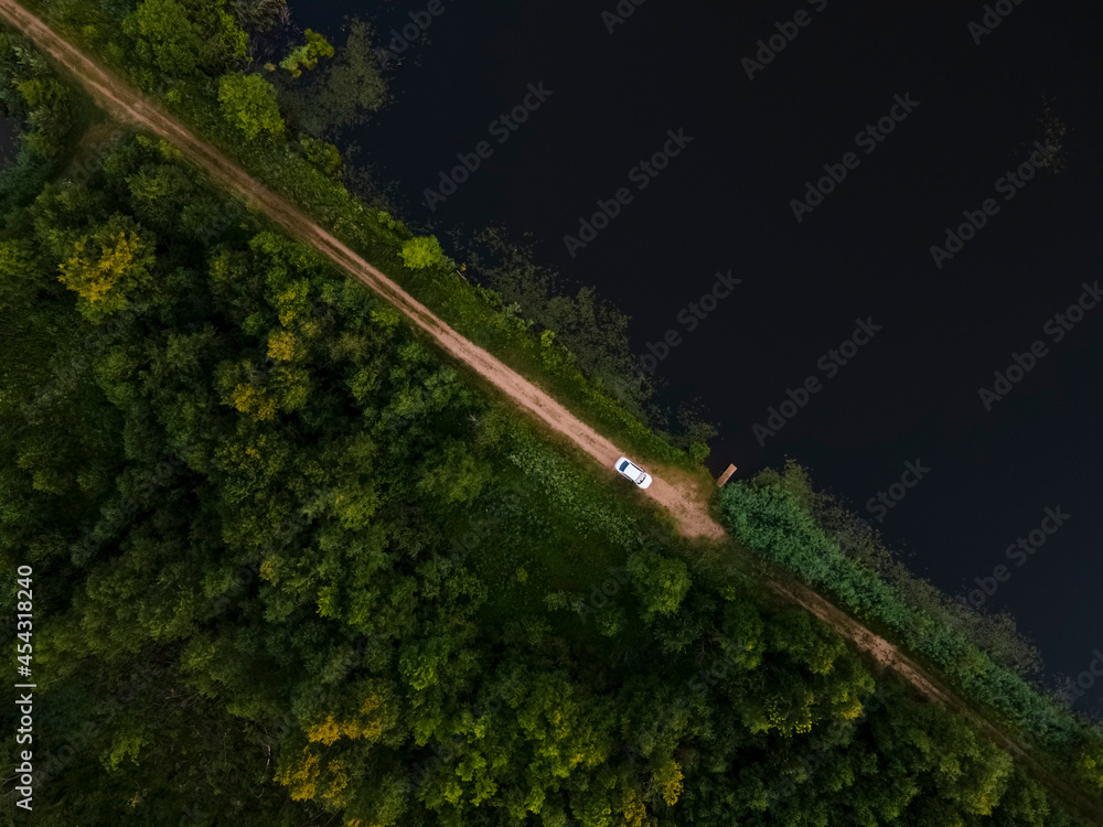 Forest and Lake aerial view landscape. Between them there is a rural road on which there is a white car. Pskov region, Russia