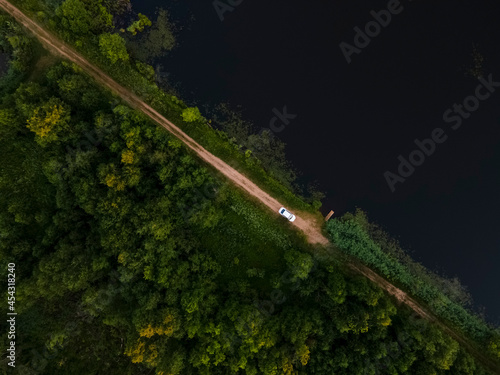 Forest and Lake aerial view landscape. Between them there is a rural road on which there is a white car. Pskov region, Russia