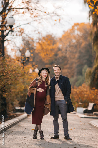 Young stylish couple in love at an autumn city park. Happy stylish man and beautiful pregnant woman in modish dress and hat posing outdoors on an autumn day
