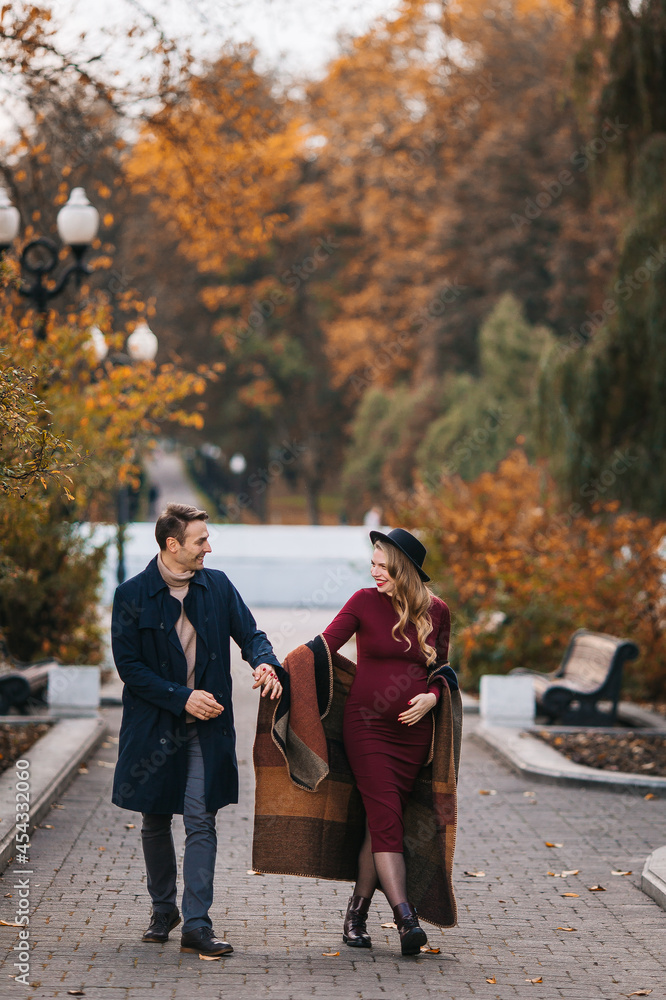 Young couple in love, happy man and his pregnant wife holding hands and walking through a park on a sunny autumn day