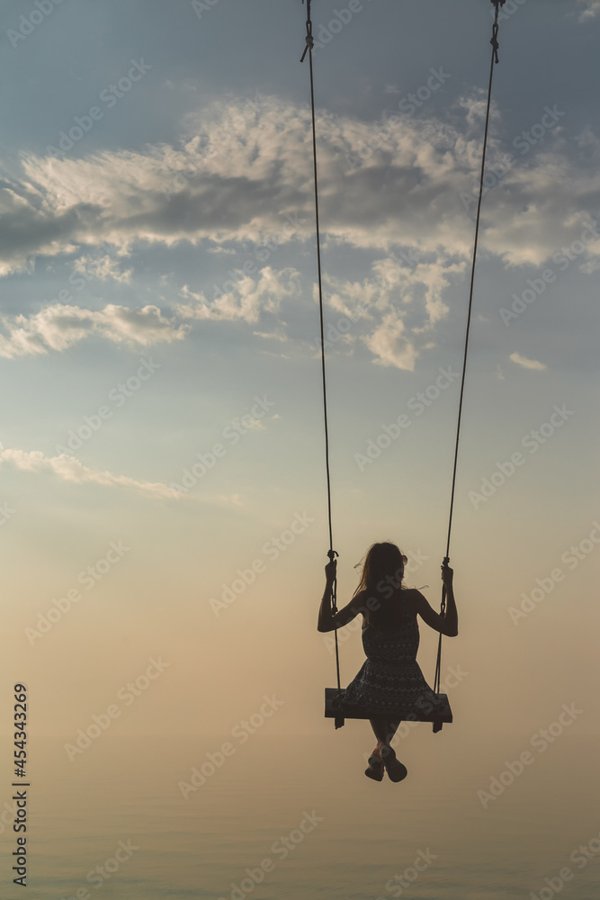Silhouette of a beautiful young girl who is swinging on a swing against the background of the sky and sea waves. Real photo without graphic editing.