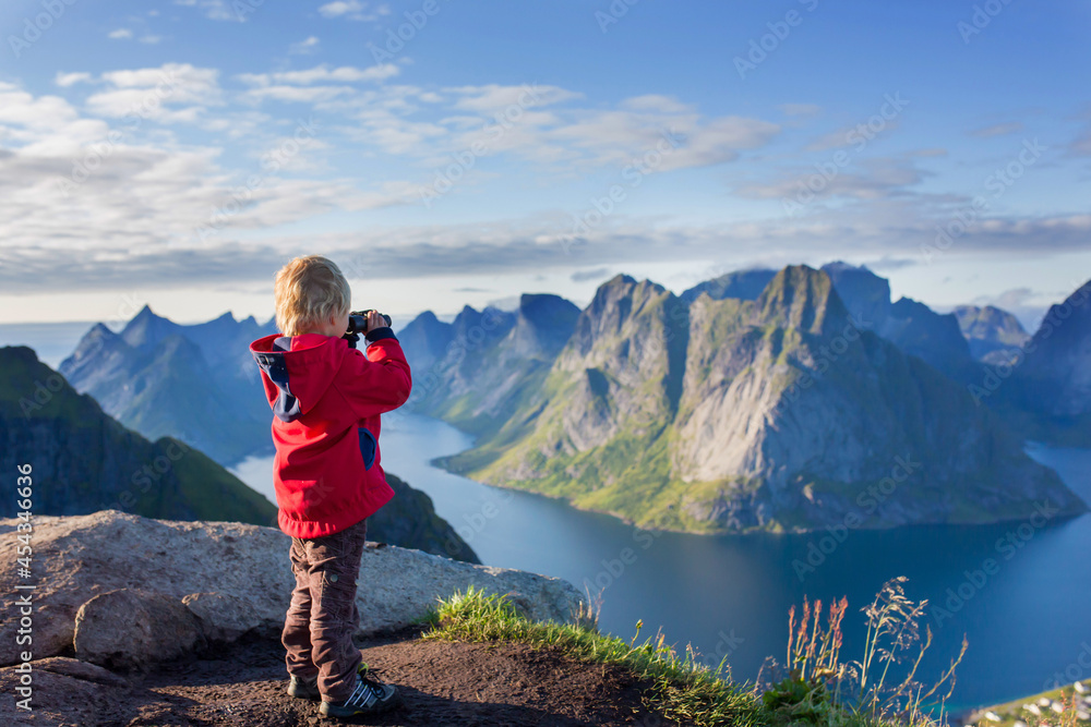 Cute child, standing on top of the mountains and looking down on Reine after climbing Reinebringen treeking path with lots of stairs, using binoculars