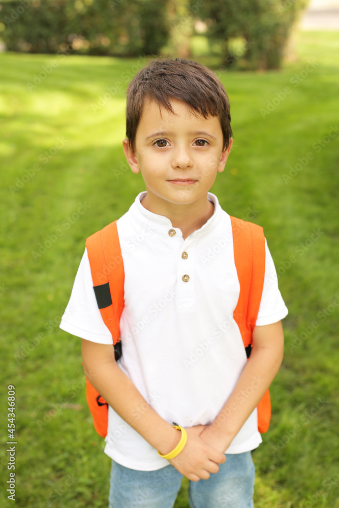 a beautiful little boy is standing in the park in a white T-shirt jeans with an orange backpack a beautiful little boy is standing in the park in a white T-shirt jeans with an orange backpack