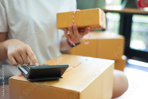Small Business Startup. Female SME Entrepreneurs Calculating Shipping Fees for Packing Boxes. Order online to prepare product boxes for sale to customers. SME business idea. © Wasan