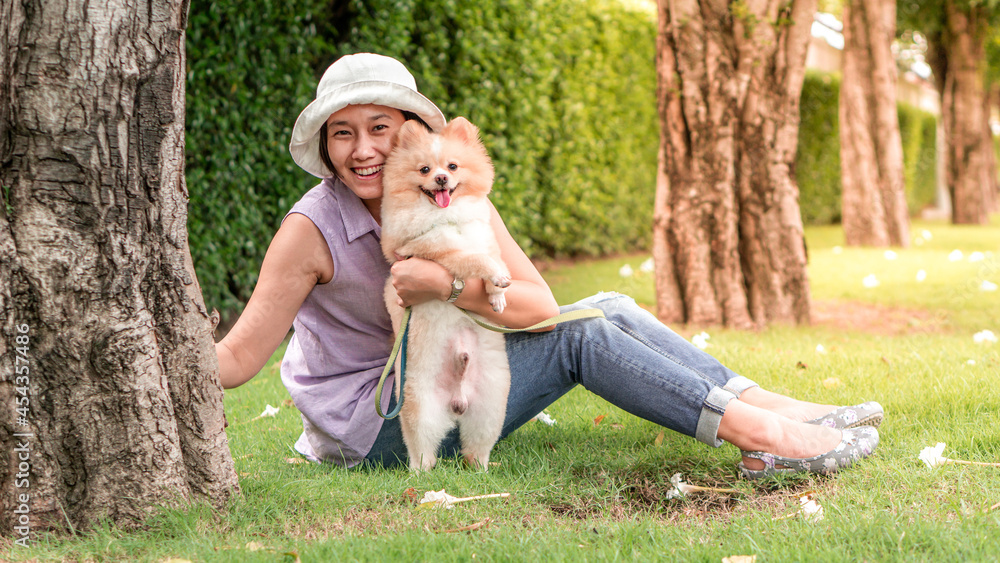 Fototapeta premium Asia women sitting on grass under a tree in the garden with lovely puppy pomeranian dog on travel tip