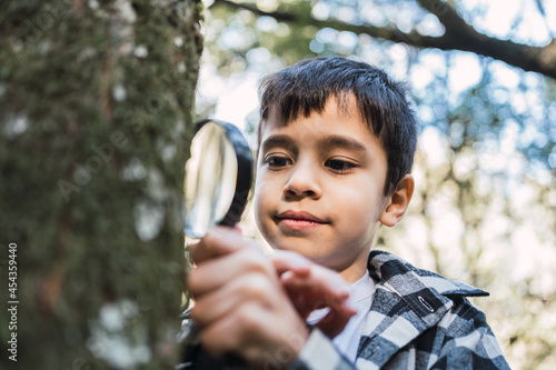 Curious ethnic boy exploring tree trunk with magnifier in woods photo