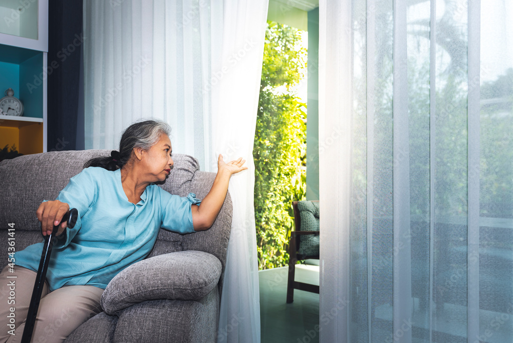 Asian elderly woman Which holds the cane to support, Sitting absent lonely on the sofa and looking outside the door, are like waiting for the child to come home, to retirement age concept. Asian elderly woman Which holds the cane to support, Sitting absent lonely on the sofa and looking outside the door, are like waiting for the child to come home, to retirement age concept.