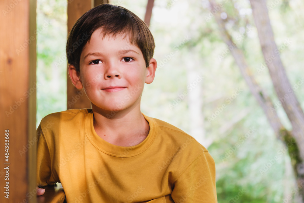 Portrait of a boy of eight years old in a yellow T-shirt. A boy with dark hair stands on the veranda of a house against the backdrop of a summer garden. Portrait of a boy of eight years old in a yellow T-shirt. A boy with dark hair stands on the veranda of a house against the backdrop of a summer garden.