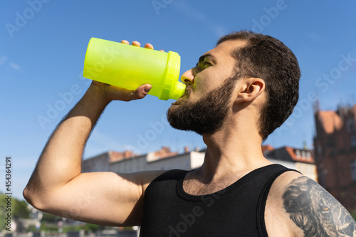 Sports man drinks water or protein cocktail outdoors. Handsome muscular guy with tattoo goes in for sports