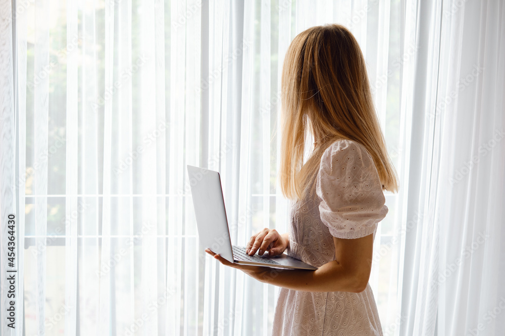 The girl works for a laptop near the window. Remote work from home or hotel. Business woman at work in the office. Business background. Home office The girl works for a laptop near the window. Remote work from home or hotel. Business woman at work in the office. Business background. Home office
