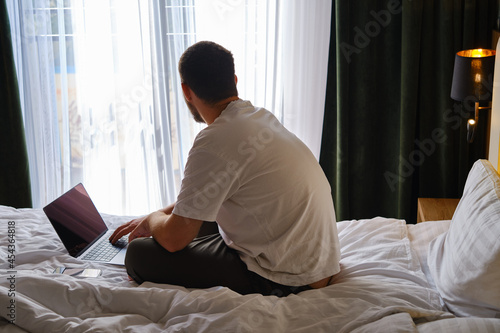 A man works with a laptop and smartphone in bed at home or in hotels. Remote work. Office at home. A man in the bedroom is typing on a laptop, distance learning for students, surfing the Internet