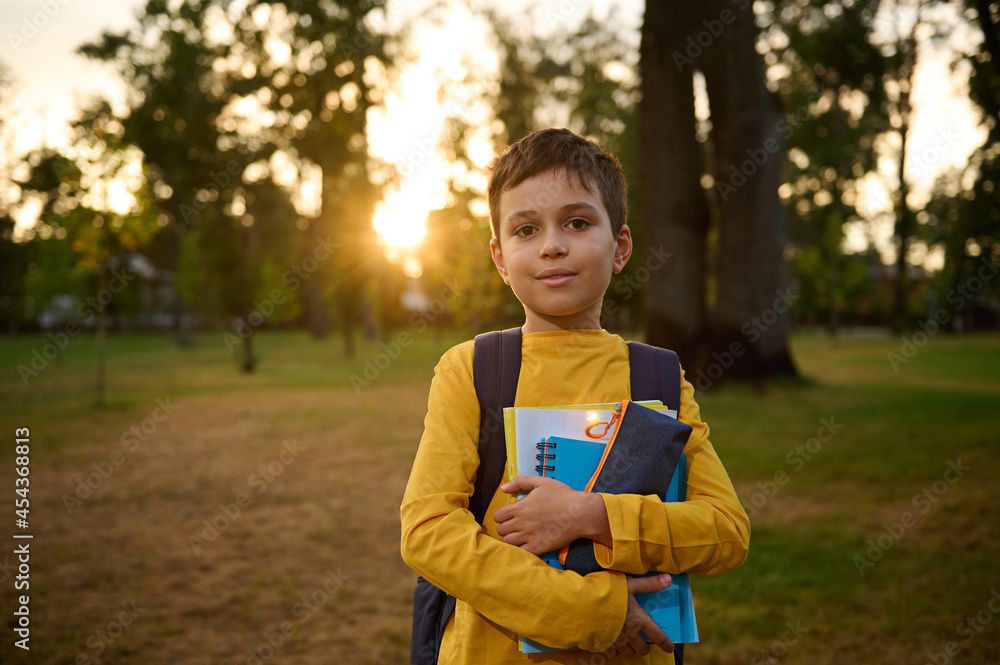 Confident portrait of a happy handsome school child boy 9 years old with backpack holding workbooks and pencil case in hands and cute smiling posing to camera against nature background at sunset Confident portrait of a happy handsome school child boy 9 years old with backpack holding workbooks and pencil case in hands and cute smiling posing to camera against nature background at sunset