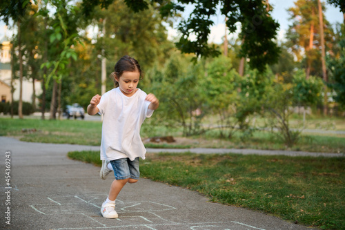 An active sporty 4 years old baby girl plays hopscotch , takes turns jumping over the squares marked on the ground. Street children's games in classics. photo