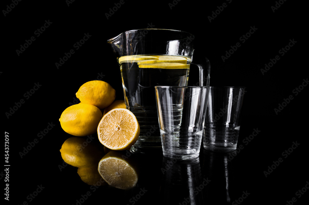 Jug or pitcher of water with lemons and two empty glasses, on black background.