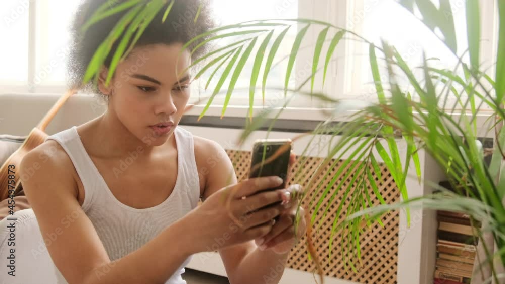 Young african american woman takes pictures photographing of a dry house plant uses a smart phone. Shares indoor plant leaves care in the mobile phone application. Young african american woman takes pictures photographing of a dry house plant uses a smart phone. Shares indoor plant leaves care in the mobile phone application.