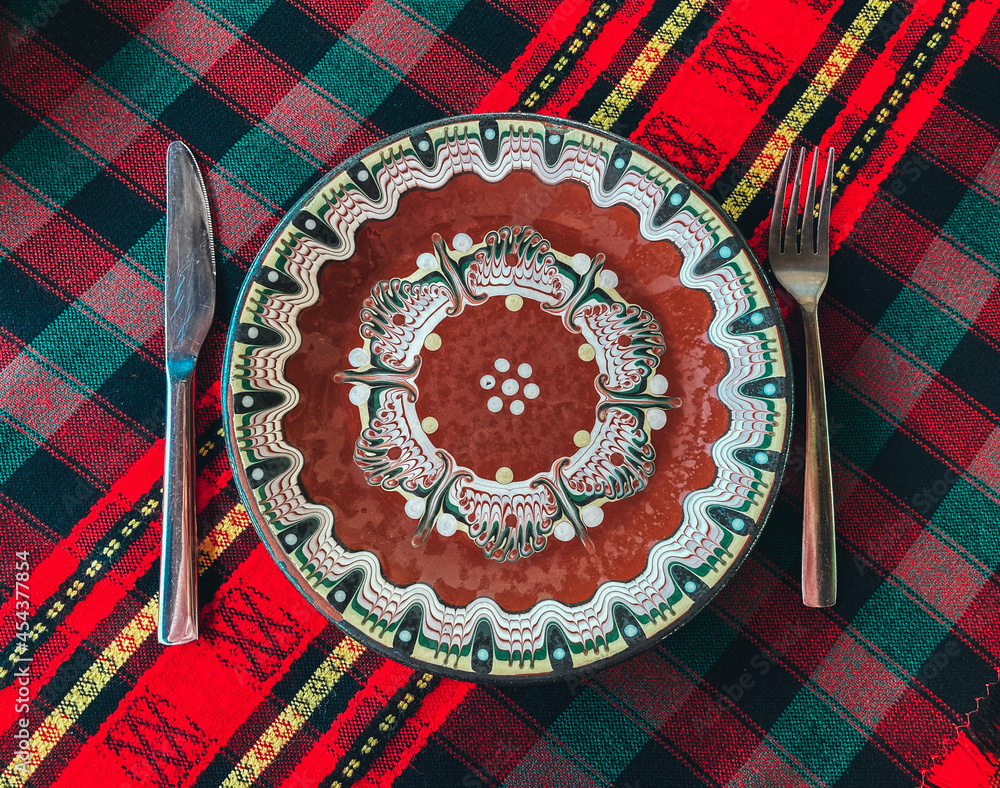 Traditional Bulgarian decorated ceramic empty plate on a red cloth, restaurant table. Fork and knife.