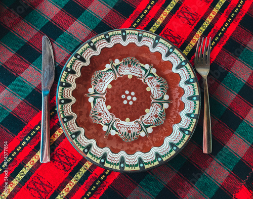 Traditional Bulgarian decorated ceramic empty plate on a red cloth, restaurant table. Fork and knife.