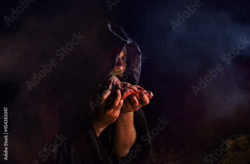 A crazy man in a black hood eats raw meat in a dark street photo