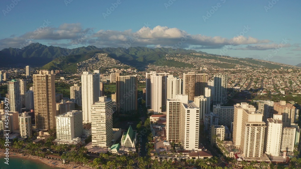 Naklejka premium Panning aerial drone slowly flying over a colorful Honolulu Skyline while Sunset in Oahu, Hawaii with Waikiki Beach Shooting from a bird's eye view. Magnificent mountains of Hawaiian island of Oahu.