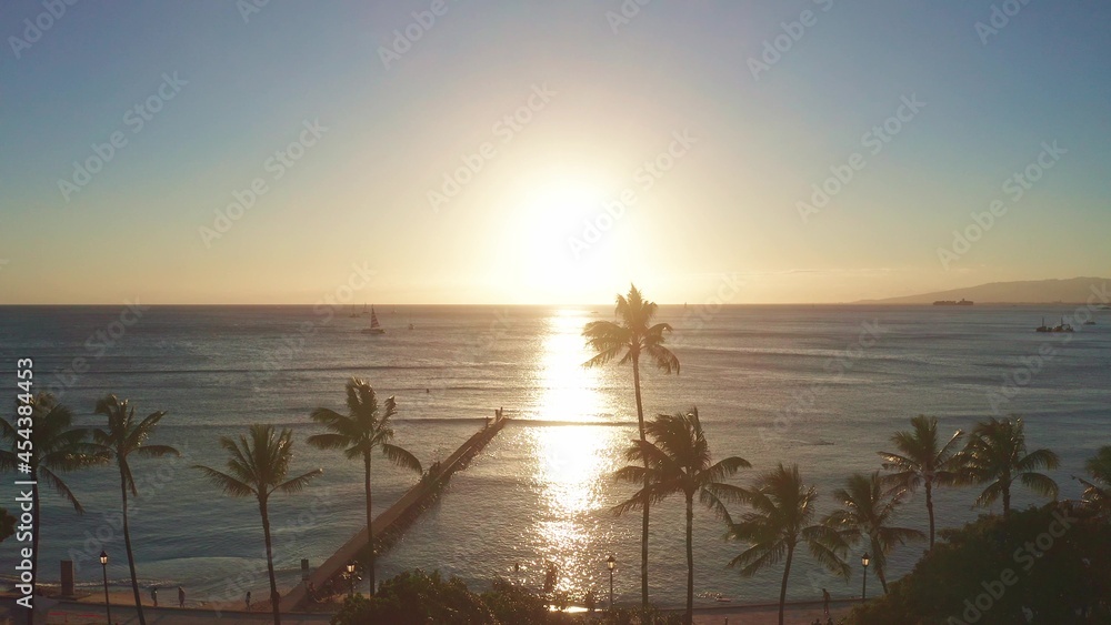 Aerial flight straight from behind between palm trees above water in ocean against colorful sunset. Panning aerial drone slowly flying over a colorful while Sunset in Oahu, Hawaii with Waikiki Beach.