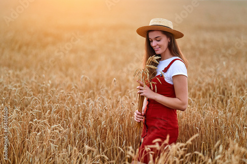 Happy smiling attractive cute free young female wearing straw hat and denim overall standing alone in golden yellow wheat field and enjoying beautiful freedom moment life at summertime