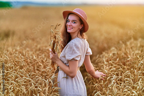 Portrait of happy smiling attractive cute free young female wearing hat and dress standing in golden yellow wheat field and enjoying beautiful freedom moment life at summertime