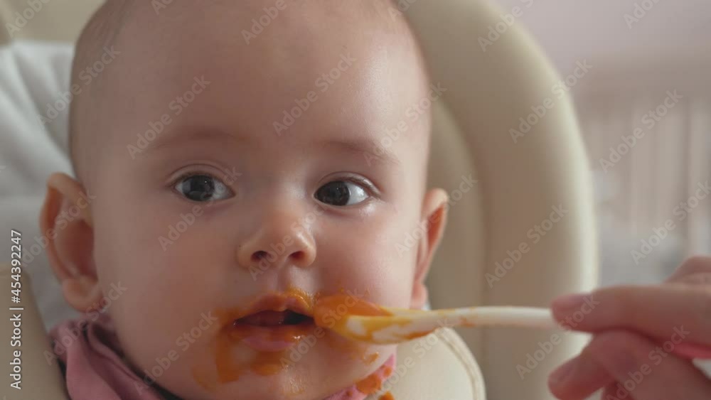 Mom's hand feeds the newborn with pumpkin puree, and lets him hold a spoon on his own Mom's hand feeds the newborn with pumpkin puree, and lets him hold a spoon on his own