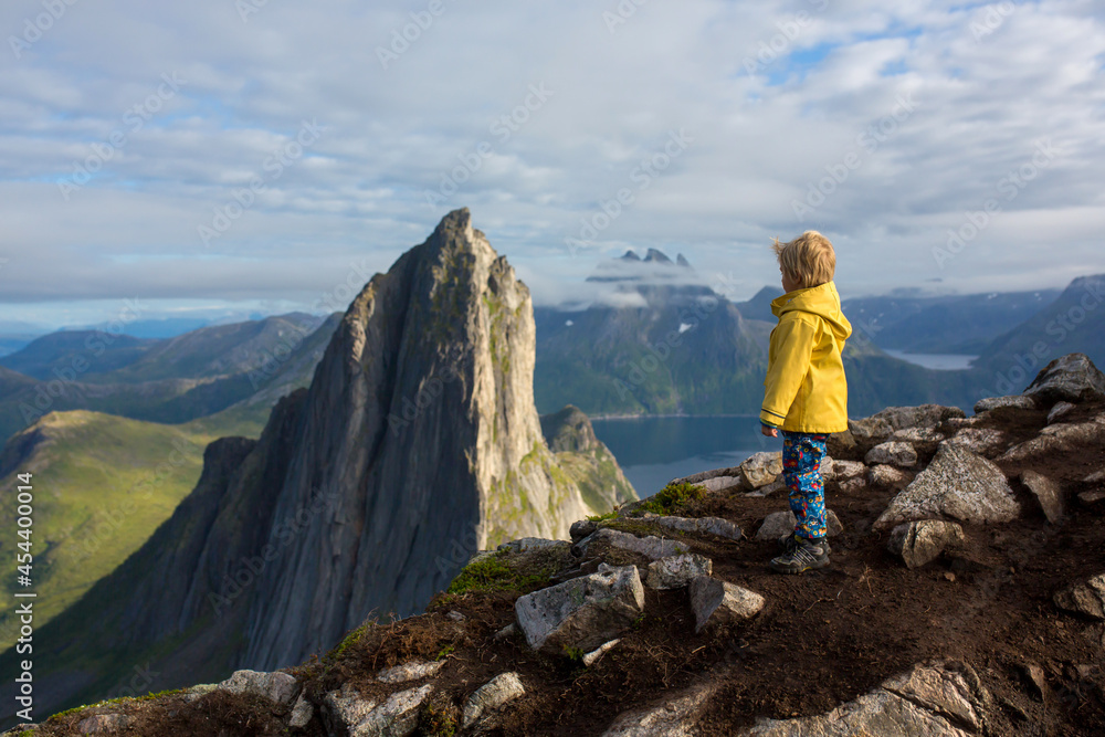 Cute toddler child in yellow raincoat, standing backwards on a rock and looking over Segla mountain on Senja island, North Norway. Amazing, beautiful landscape and splendid nature