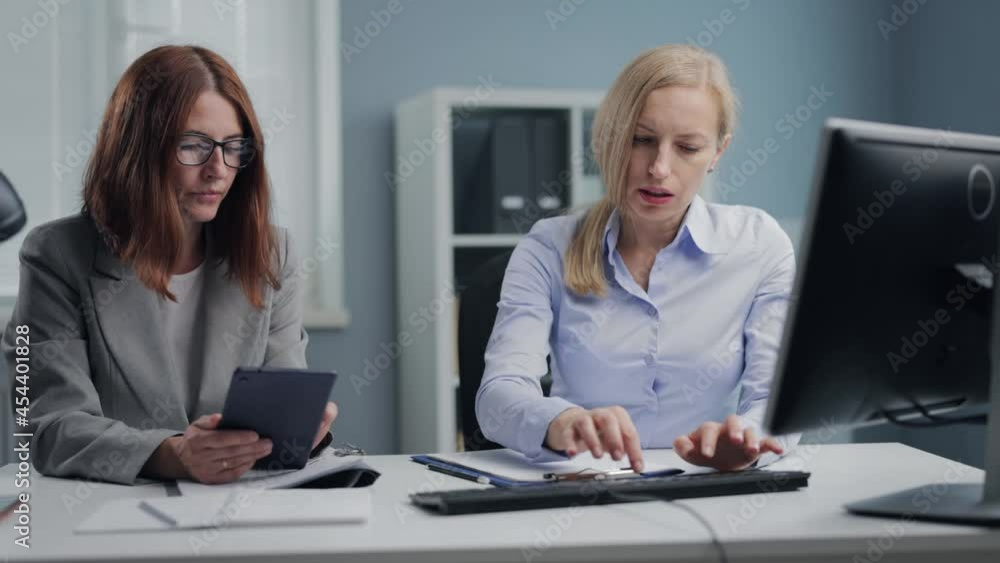 Confident business women working on computer and tablet during meeting at bright office. Two caucasian female sitting at desk and discussing working project. Cooperation concept. Confident business women working on computer and tablet during meeting at bright office. Two caucasian female sitting at desk and discussing working project. Cooperation concept.