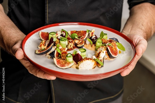 People at work. The chef's hands are holding a plate with summer salad with ripe figs, goat chevre cheese, fresh cucumber roll, beetroot and mustard leaves with tomato sauce. Restaurant dish