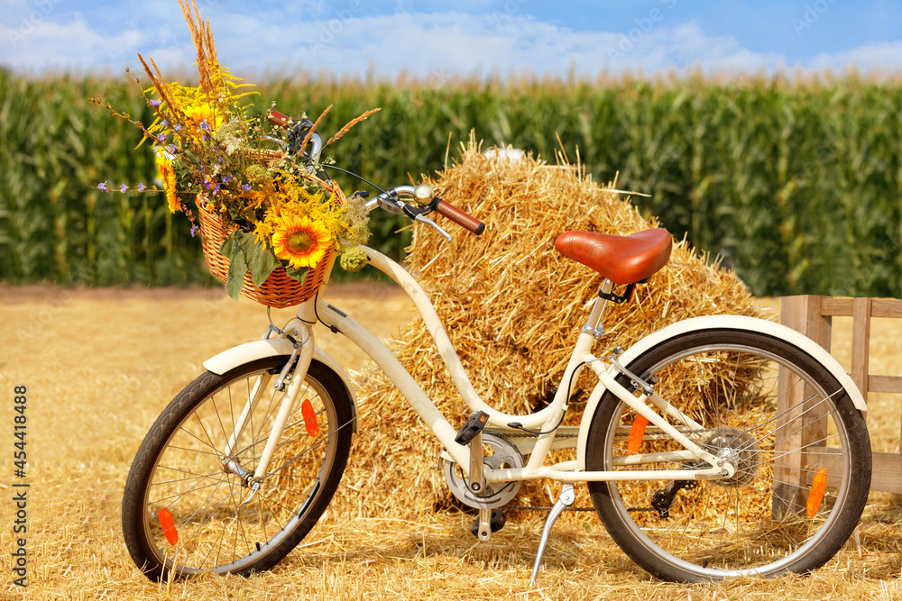 Naklejka premium Ladies bicycle with a basket, a bouquet of wild flowers in front on the handlebars against the background of a heap of straw and a corn field in blur.