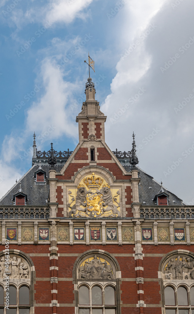 Amsterdam, Netherlands - August 14, 2021: Frontal monumental sculpted red stone middle facade with tower and coat of arms of Centraal railway station under blue cloudscape.